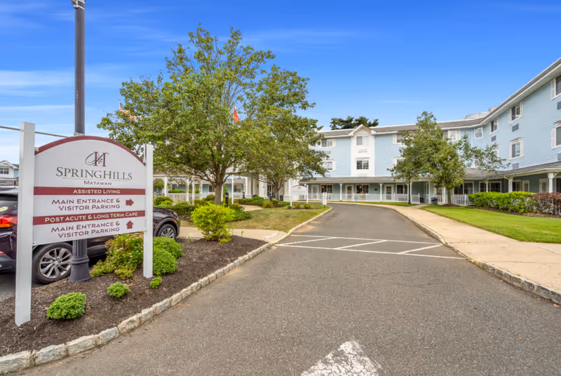 Exterior view of Spring Hills Matawan senior living facility showing a driveway leading to the main entrance. There is a sign on the left indicating directions for assisted living, main entrance and visitor parking, and post acute & long term care. The building is light blue with white trim, surrounded by green trees and landscaping under a clear blue sky.