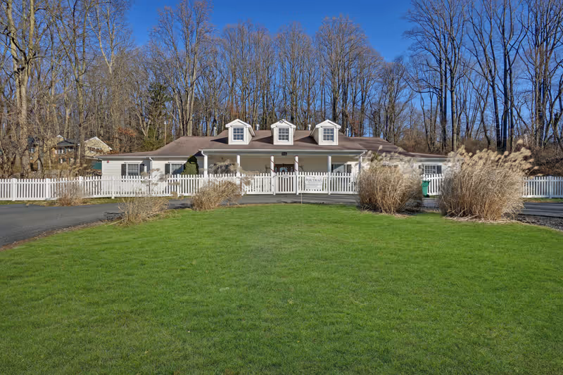 A single-story building with a brown roof and three dormer windows, surrounded by a white picket fence. The building is set against a backdrop of leafless trees under a clear blue sky. In front of the building is a well-maintained green lawn with some ornamental grasses near the driveway.