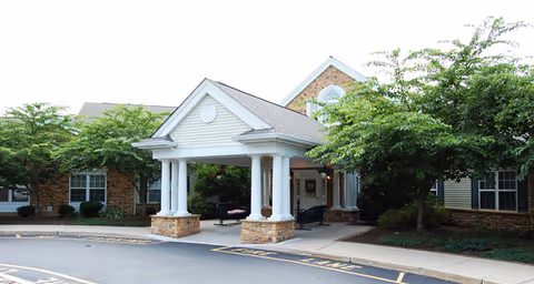 Front exterior view of Brookdale Hamilton facility showing a covered entrance with white columns, brick and siding walls, surrounded by trees and greenery.