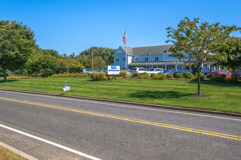 Exterior view of Complete Care at Holmdel facility with a parking lot filled with cars, a green lawn with trees, an American flag on a flagpole, and a sign displaying the facility's name.