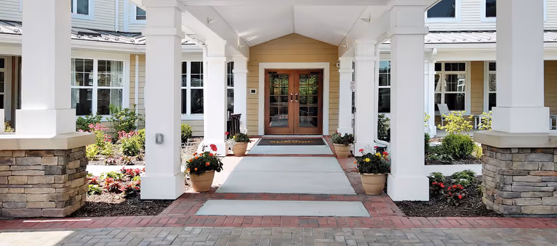 Covered entrance with white columns, potted flowers, brick walkway, and double wooden doors of a senior living building.
