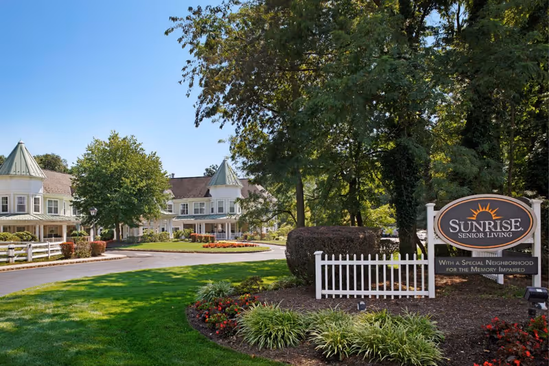 Exterior view of Sunrise Senior Living facility with a large sign in the foreground and a two-story building with green roofs and white fences surrounded by trees and landscaping under a clear blue sky.