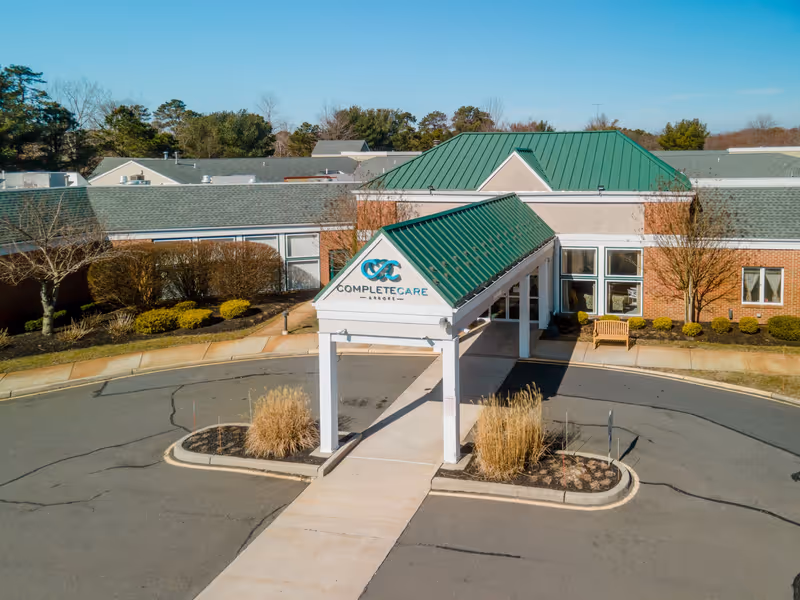 Exterior front entrance of Complete Care at Arbors facility with a covered driveway, green roof, brick walls, and landscaped bushes and trees under a clear blue sky.