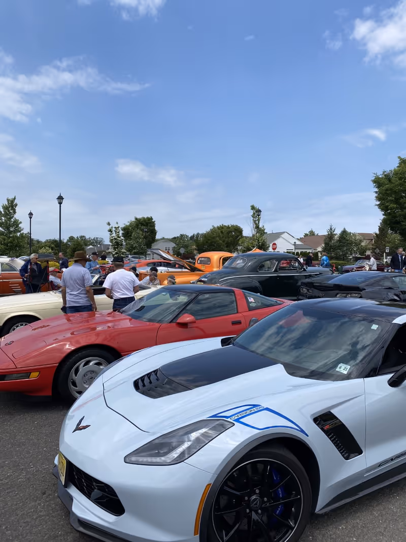 A parking lot filled with various classic and modern cars, including a white sports car with black and blue detailing in the foreground and a red sports car behind it. Several people are walking around and looking at the cars under a partly cloudy blue sky.