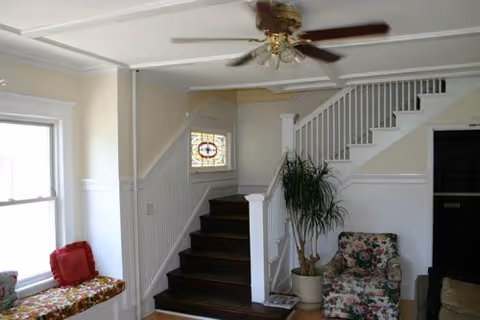 Interior view of a living area with a staircase leading upstairs. There is a ceiling fan with lights, a window with a stained glass panel, a potted plant, a floral upholstered chair, and a cushioned bench by a large window.