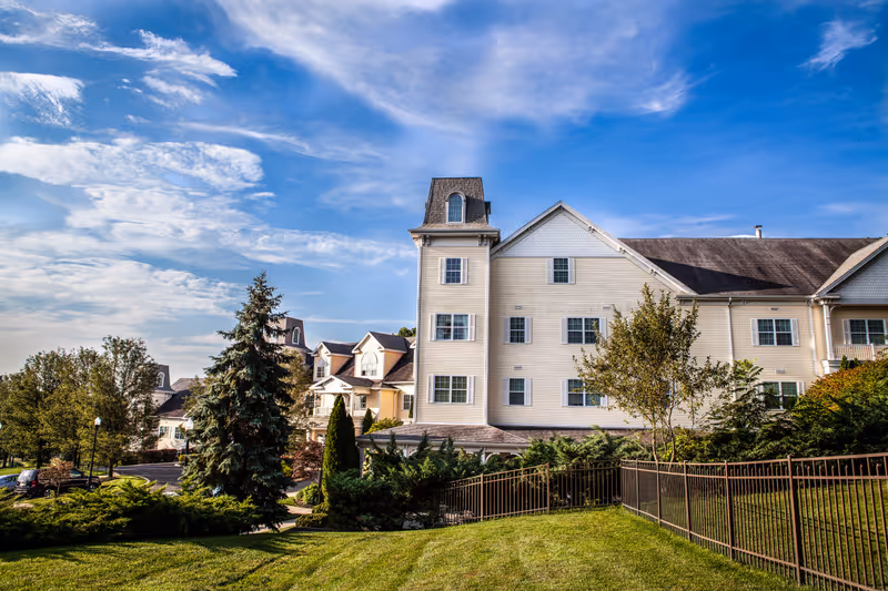 Exterior view of a multi-story senior living facility building with beige siding, multiple windows, and a tower-like structure. The building is surrounded by well-maintained green lawns, trees, and shrubs under a partly cloudy blue sky.
