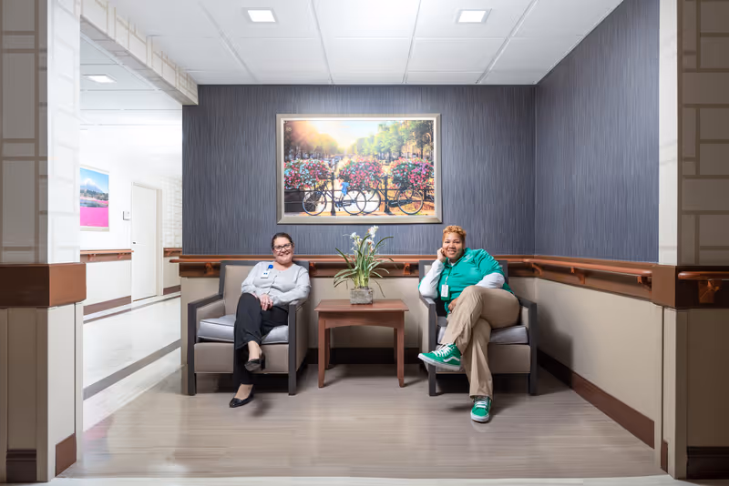 Two women sitting in armchairs in a hallway seating area of a healthcare facility. There is a small wooden table with a plant between them and a framed picture of bicycles with flower baskets on the wall behind them. The hallway has light-colored floors and walls with handrails.