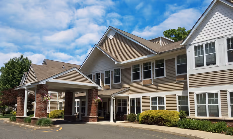 Exterior view of a senior living facility building with beige siding and white trim, featuring a covered entrance supported by brick columns, surrounded by greenery and under a partly cloudy blue sky.