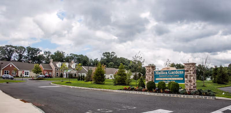 Exterior view of Mullica Gardens Assisted Living Center showing a paved driveway, landscaped greenery with small trees and bushes, and a large sign with the facility's name. The building is visible in the background under a cloudy sky.