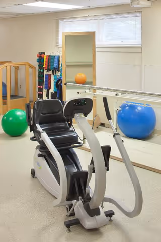 A small exercise room with a recumbent exercise bike in the foreground. Behind it, there are colorful dumbbells on a rack, resistance bands hanging, a large mirror, and two exercise balls, one green and one blue. The room has light-colored walls and a window with blinds near the ceiling.