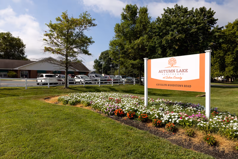 A landscaped lawn and flower bed in front of a large sign for Autumn Lake Healthcare at Salem County with the facility building and parked cars in the background.