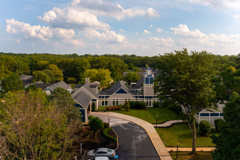 Aerial view of Harrogate Retirement Community showing multiple buildings with gray roofs surrounded by green trees and lawns under a partly cloudy sky. A curved driveway leads to the main building with a clock tower.