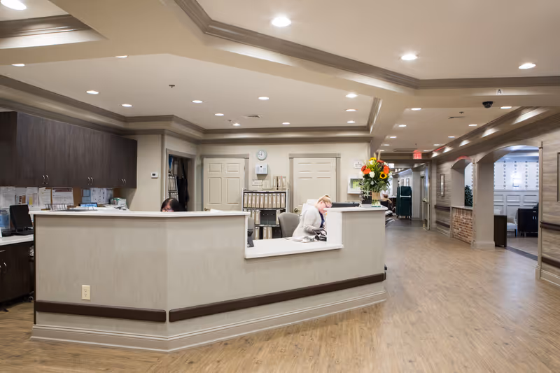 Reception area inside CareOne at Wall facility with a curved front desk, two staff members working behind the desk, wood flooring, overhead recessed lighting, and a hallway leading to other rooms. There is a vase with colorful flowers on the desk and filing cabinets in the background.