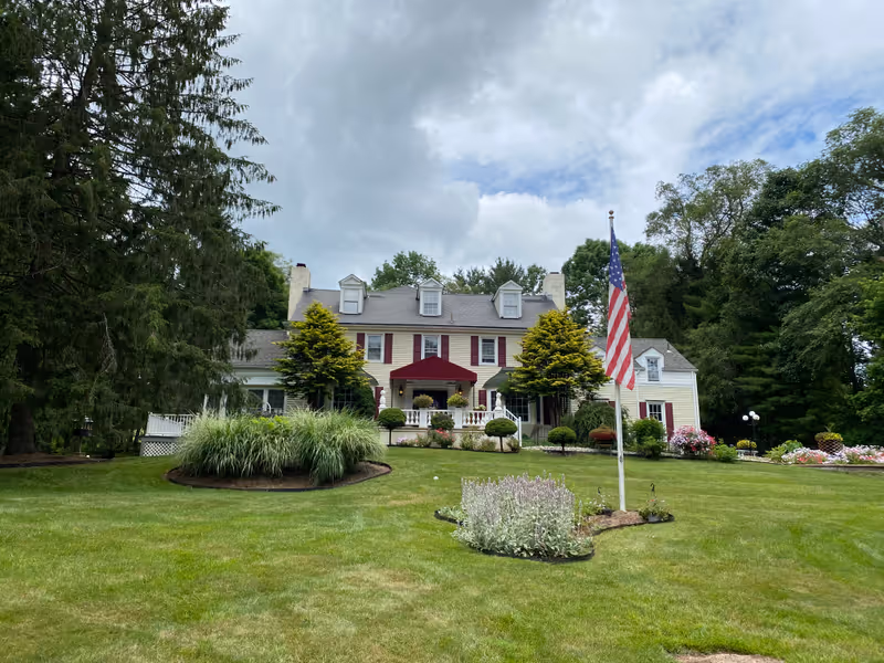 Front exterior view of a large, two-story colonial-style building with white siding and red shutters, surrounded by well-maintained green lawns, shrubs, and trees. An American flag is displayed on a flagpole in the front yard under a partly cloudy sky.