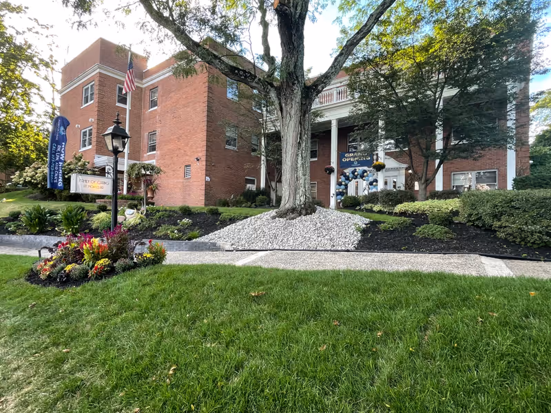 Front exterior of a red-brick senior living building with columns, landscaped lawn, and a decorated entrance.