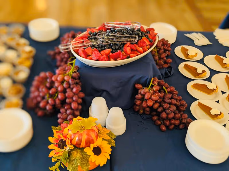 A buffet table with a large bowl of mixed berries including strawberries and blueberries, surrounded by bunches of red grapes. On the right side, there are several plates with slices of pumpkin pie topped with whipped cream. The table is covered with a dark blue tablecloth and decorated with a small autumn-themed centerpiece featuring a pumpkin and sunflowers.