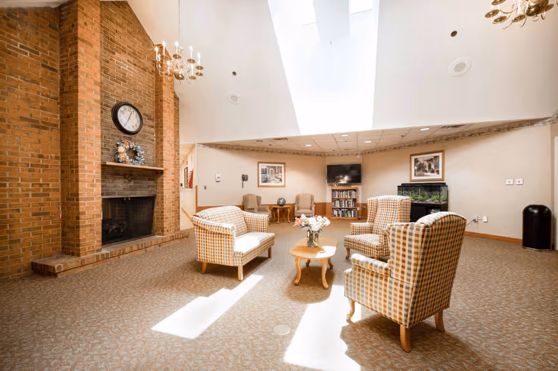 A bright and spacious common area with a high ceiling and skylight. The room features a brick fireplace with a clock and wreath above it, a set of four plaid upholstered chairs arranged around a wooden coffee table with a flower vase, two armchairs with a small table in the background, a television mounted on the wall above a bookshelf, and an aquarium. The walls are decorated with framed pictures and the floor is carpeted.
