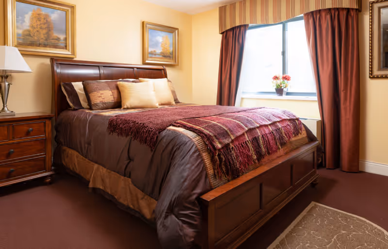 A cozy bedroom with a wooden bed frame, a neatly made bed with brown and burgundy bedding, and several pillows. There is a wooden nightstand with a lamp on the left side of the bed. The room has beige walls with two framed paintings of autumn trees. A window with burgundy curtains and a valance lets in natural light, and a small potted plant sits on the windowsill. A beige rug with a subtle pattern is partially visible on the floor.