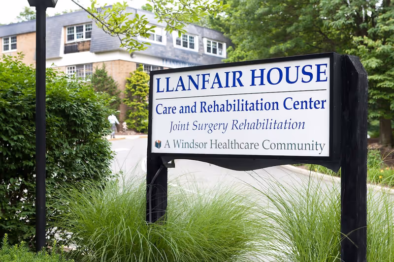 Outdoor view of a sign for Llanfair House Care and Rehabilitation Center surrounded by greenery, with part of the building visible in the background.