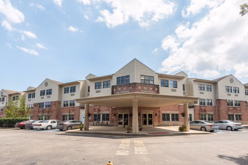 Exterior view of a three-story senior living facility building with a covered entrance supported by columns. Several cars are parked in front of the building under a partly cloudy sky.