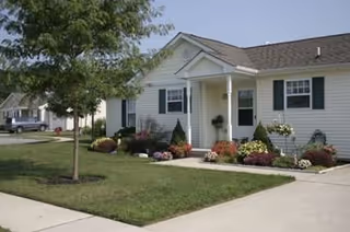 Single-story residential building with white siding, black shutters, and a small front porch surrounded by well-maintained landscaping including flowers and shrubs. A tree and a sidewalk are visible in the foreground.