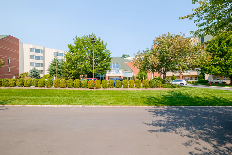 Front exterior of a multi-story senior living building with a green lawn, trimmed shrubs, trees, and a central glass entrance.
