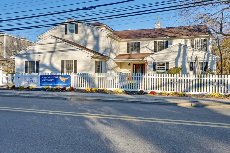 Exterior front view of a two-story white house with a brown roof, surrounded by a white picket fence and colorful flowers along the fence line. A banner on the fence reads 'Fox Trail Memory Care Living' with a call to schedule a visit and contact information. The house is located along a paved street with double yellow lines.