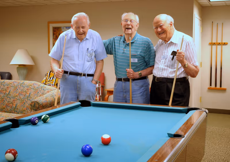 Three elderly men standing around a pool table indoors, each holding a pool cue and smiling. The room has a floral patterned couch, a lamp, a framed picture on the wall, and a rack with additional pool cues.