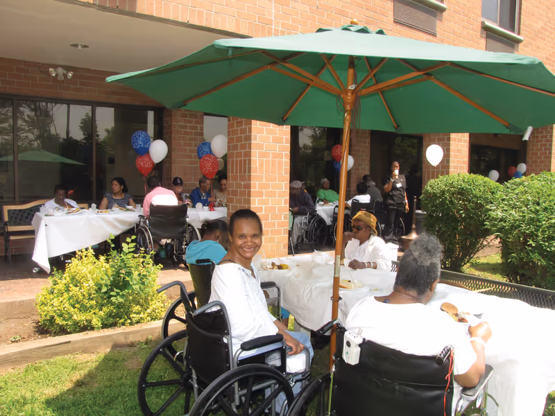 Outdoor gathering at New Community Extended Care with elderly residents, some in wheelchairs, seated at tables covered with white tablecloths under a large green umbrella. Red, white, and blue balloons are tied to the tables and the brick building is visible in the background.