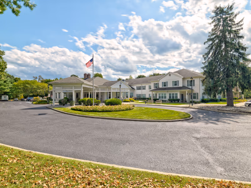Exterior view of a senior living facility named The Residence at Voorhees, showing a large two-story white building with a covered entrance, an American flag on a flagpole, a circular driveway, and landscaped greenery under a partly cloudy sky.
