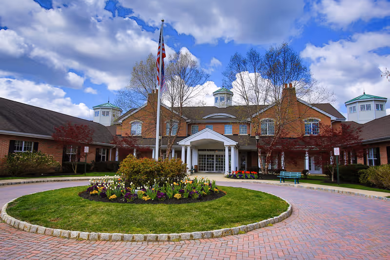 Front exterior view of a senior living facility with a circular driveway, a flagpole with an American flag, landscaped flower beds, and a brick building with white columns and multiple windows under a partly cloudy sky.