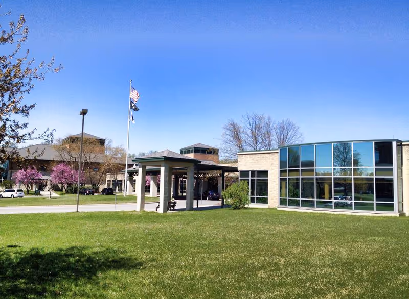 Exterior view of Elmwood Hills Healthcare Center showing a modern building with large windows, a covered entrance, flagpoles with flags, green lawn, and trees with pink blossoms under a clear blue sky.