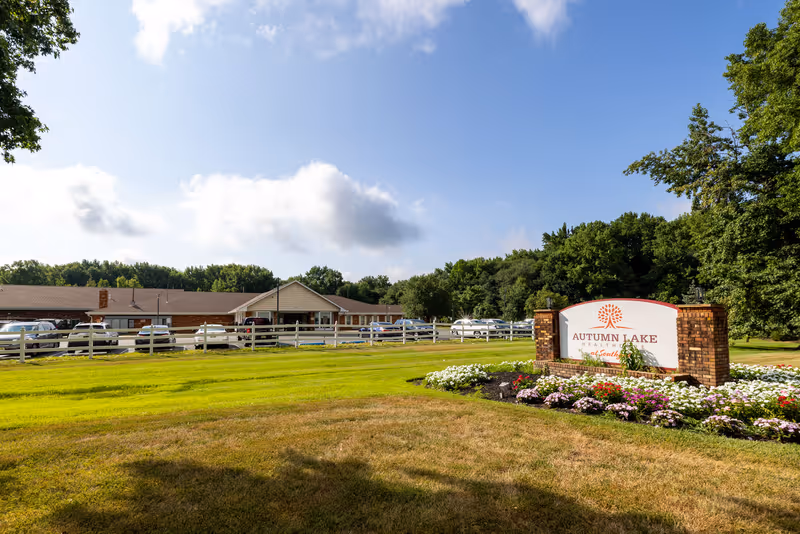 Exterior view of Autumn Lake Healthcare at Southgate facility with a large green lawn, a white fence, several parked cars, and a clear blue sky with some clouds. A brick sign with flowers around it displays the facility name.