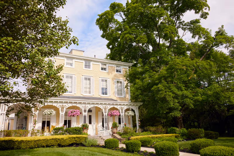 A large, three-story yellow building with white trim and decorative porch railings surrounded by lush green trees and neatly trimmed bushes under a partly cloudy sky.