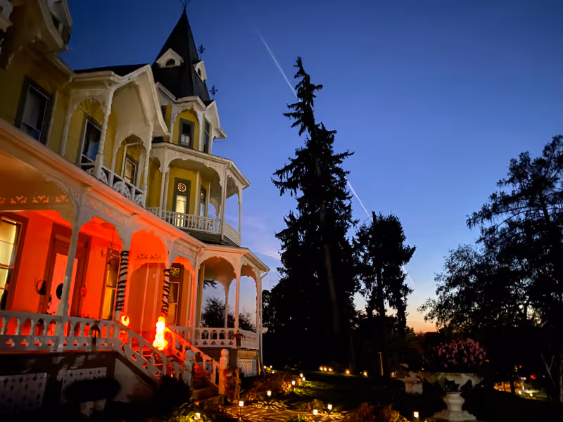 A large Victorian-style house with a wraparound porch illuminated by orange lights at dusk, surrounded by tall trees and a garden with pathway lights.