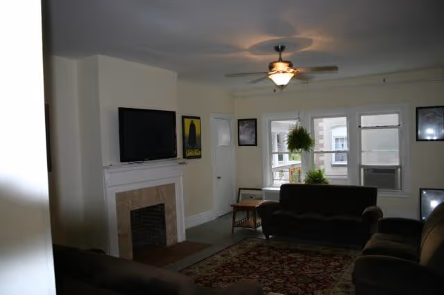 A living room with beige walls and a ceiling fan with light. The room features a fireplace with a mounted flat-screen TV above it. There are three dark-colored sofas arranged around a patterned area rug. A small wooden side table is placed near a window with a hanging plant and an air conditioning unit. Several framed pictures hang on the walls.
