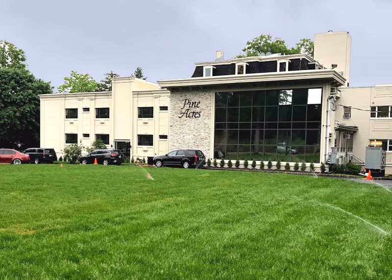 Front exterior of the Pine Acres building with a large green lawn, parked cars, and a wall of reflective windows.