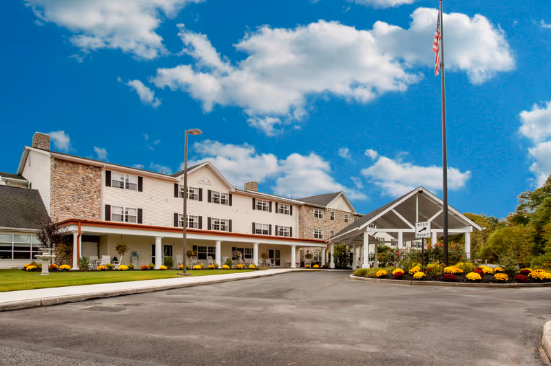 Exterior view of a senior living facility building with a covered entrance, stone and white siding walls, multiple windows, a flagpole with an American flag, and landscaped flower beds under a partly cloudy blue sky.
