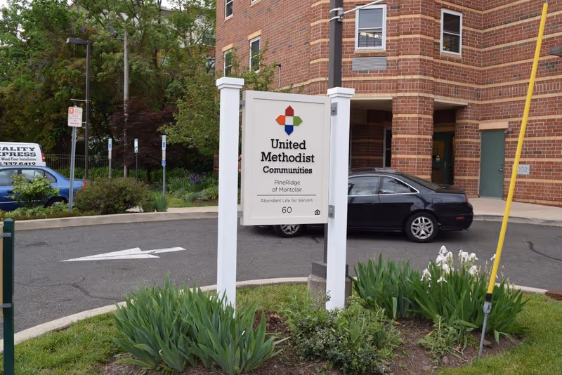 White sign reading 'United Methodist Communities' stands in front of a brick senior living building with parked cars and landscaped greenery.