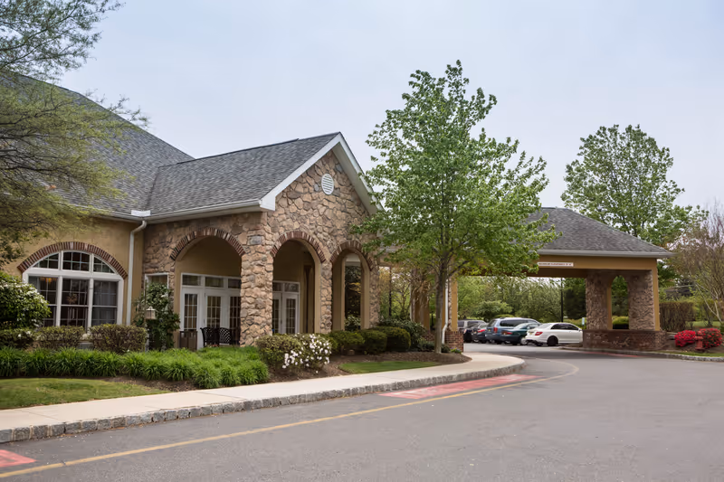 Exterior view of a senior living facility building with stone and brick accents, arched entryways, and a covered driveway. There are several trees and shrubs around the building, and cars are parked in the parking lot nearby under a cloudy sky.