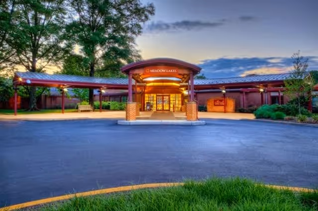 Front exterior view of Meadow Lakes facility at dusk with a covered entrance, brick pillars, and surrounding trees and greenery.
