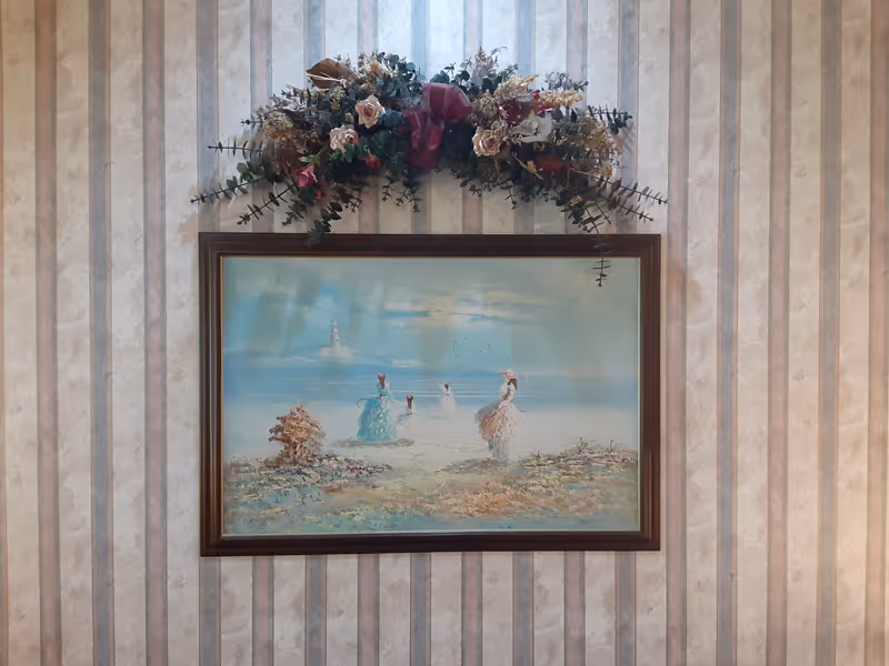 A framed painting hanging on a vertically striped wallpapered wall. The painting depicts a serene beach scene with four women in vintage dresses walking along the shore, with a lighthouse in the background. Above the painting is a decorative floral arrangement with various flowers and greenery.