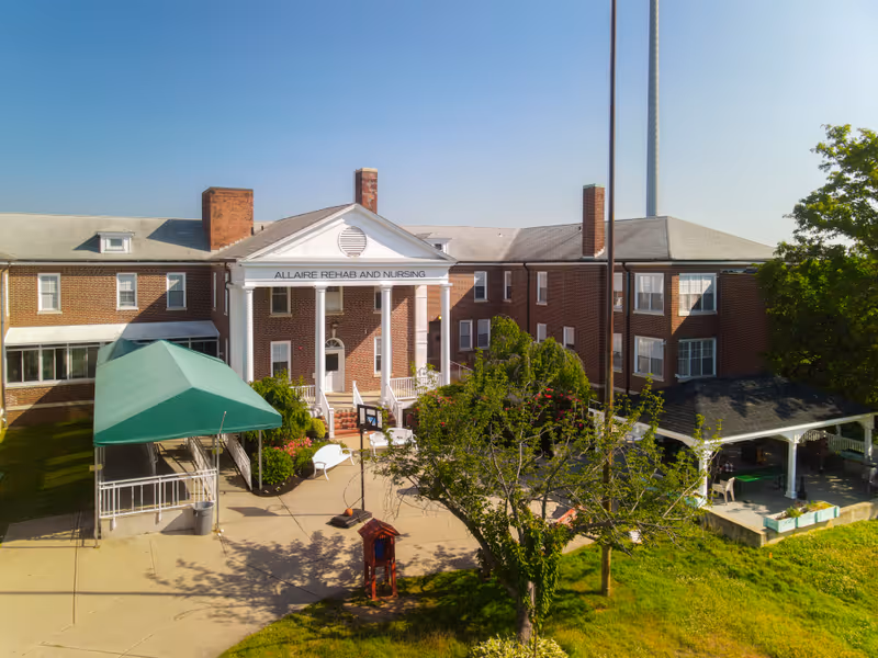 Exterior view of Allaire Rehab and Nursing facility showing a large brick building with white columns at the entrance, a green canopy over a ramp, a basketball hoop, trees, and a covered patio area on a sunny day.
