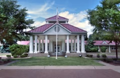 Front entrance of a senior living facility with white columns, a central flagpole, and landscaped grounds.