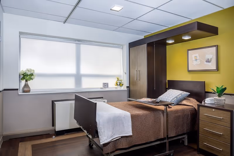 Private resident bedroom with a single bed, over-bed table, wardrobe, bedside drawers, and a large window with a blind.