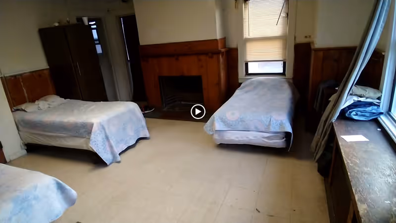 A simple bedroom with three single beds covered with light blue patterned bedspreads. The room has wood paneling on the walls, a fireplace in the center, a wardrobe in the corner, and a window with blinds partially drawn. There is a wooden ledge under the window with some folded clothes and a piece of paper on it.