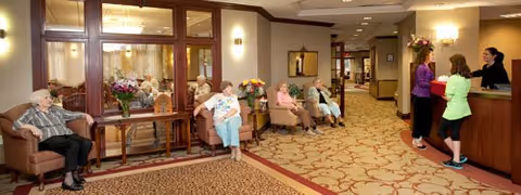 A senior living facility lobby area with elderly residents seated on chairs along the wall and a reception desk where two women are interacting with a receptionist. The space is warmly lit with carpeted floors and decorative flowers on tables.