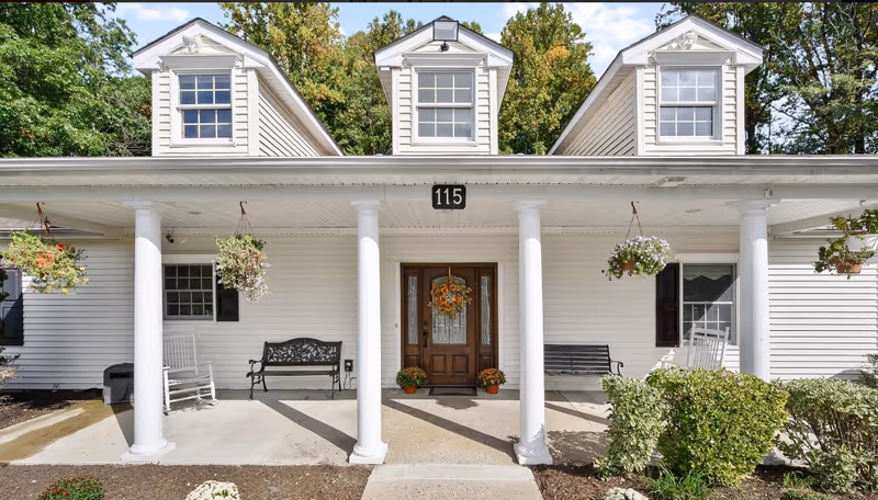 Front exterior view of a white building with three dormer windows on the roof, a covered porch supported by four white columns, hanging flower baskets, benches, rocking chairs, and a wooden door decorated with a wreath and potted plants on either side. The building number 115 is displayed above the door.