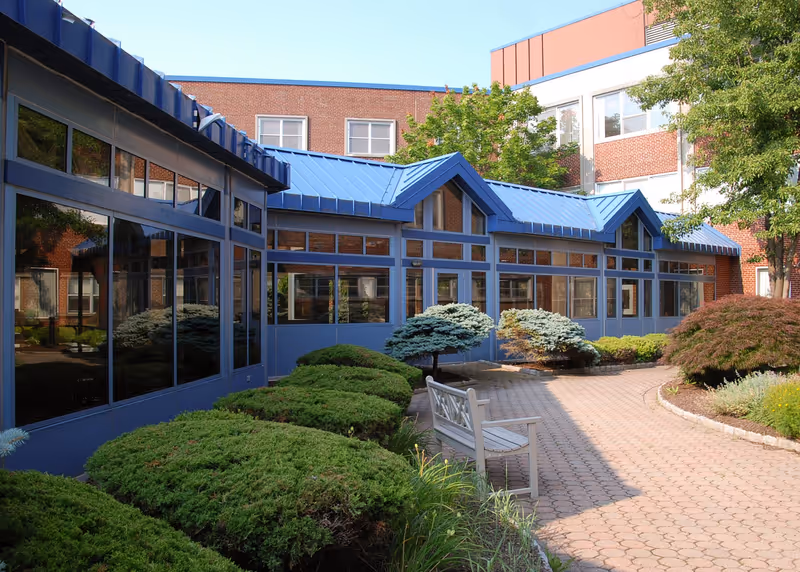 Outdoor courtyard area of a healthcare facility with a paved walkway, green bushes, small trees, and a white bench. The building has blue metal roofing and large windows reflecting the greenery.