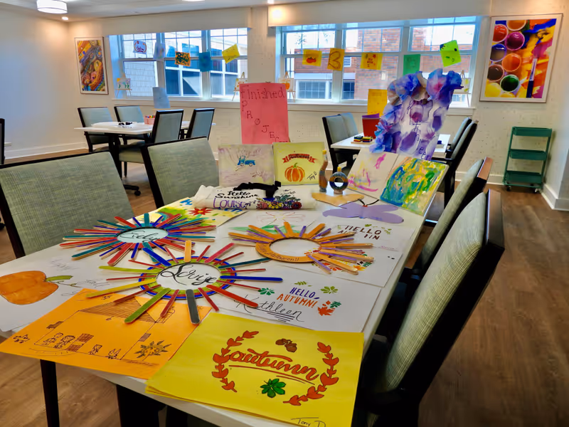 A bright activity room with tables and chairs arranged for crafts. The main table is covered with colorful autumn-themed art projects including drawings, painted popsicle stick wreaths, and paper crafts. The walls have framed artwork and windows letting in natural light. Additional tables and chairs are visible in the background.